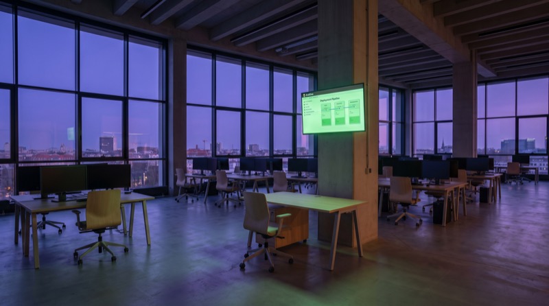 La planta de desarrollo de Pixel Spree bajo un profundo crepúsculo violeta de verano en Berlín. La oficina está vacía, con escritorios ordenados, monitores oscuros y una sola pantalla mural que muestra un bloque sólido de pipeline de Jenkins brillando en verde.